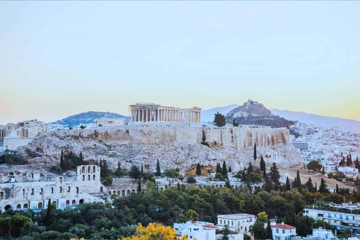 The ruins of a stone temple on a white outcropping of rock with white buildings of Athens among the trees below