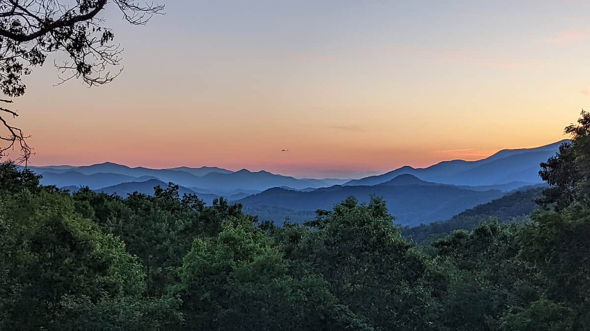 A serene landscape of mountain ranges during sunset, with layers of hills fading into the distance. The sky transitions from a soft orange near the horizon to a light blue higher up. Green trees fill the foreground, adding depth to the scenery.