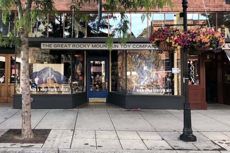 Street view of The Great Rocky Mountain Toy Company, featuring large storefront windows displaying an array of toys. The entrance is adorned with colorful hanging plants, and trees line the sidewalk in front.