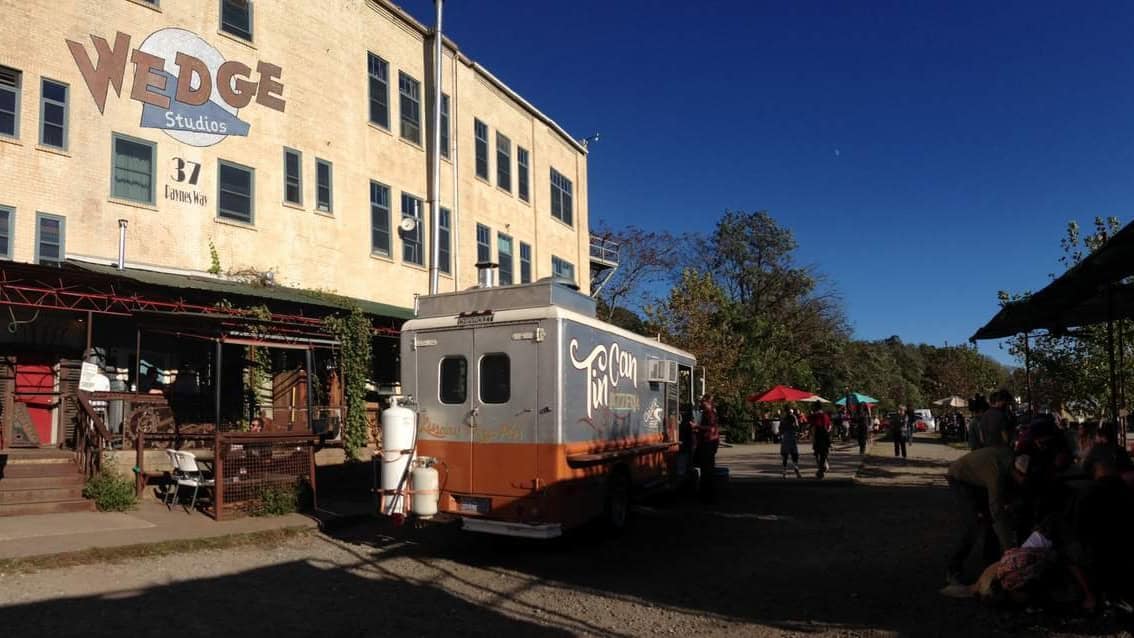 A large beige building with the " Wedge Studios" written on the side in a graphic design. A silver food truck sits in front and there are people eating at tables, typical of an Asheville brewery.