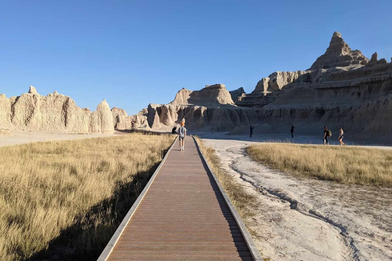 A wooden boardwalk extends straight out with a kid walking down the middle. Rock formations extend upwards on the right side