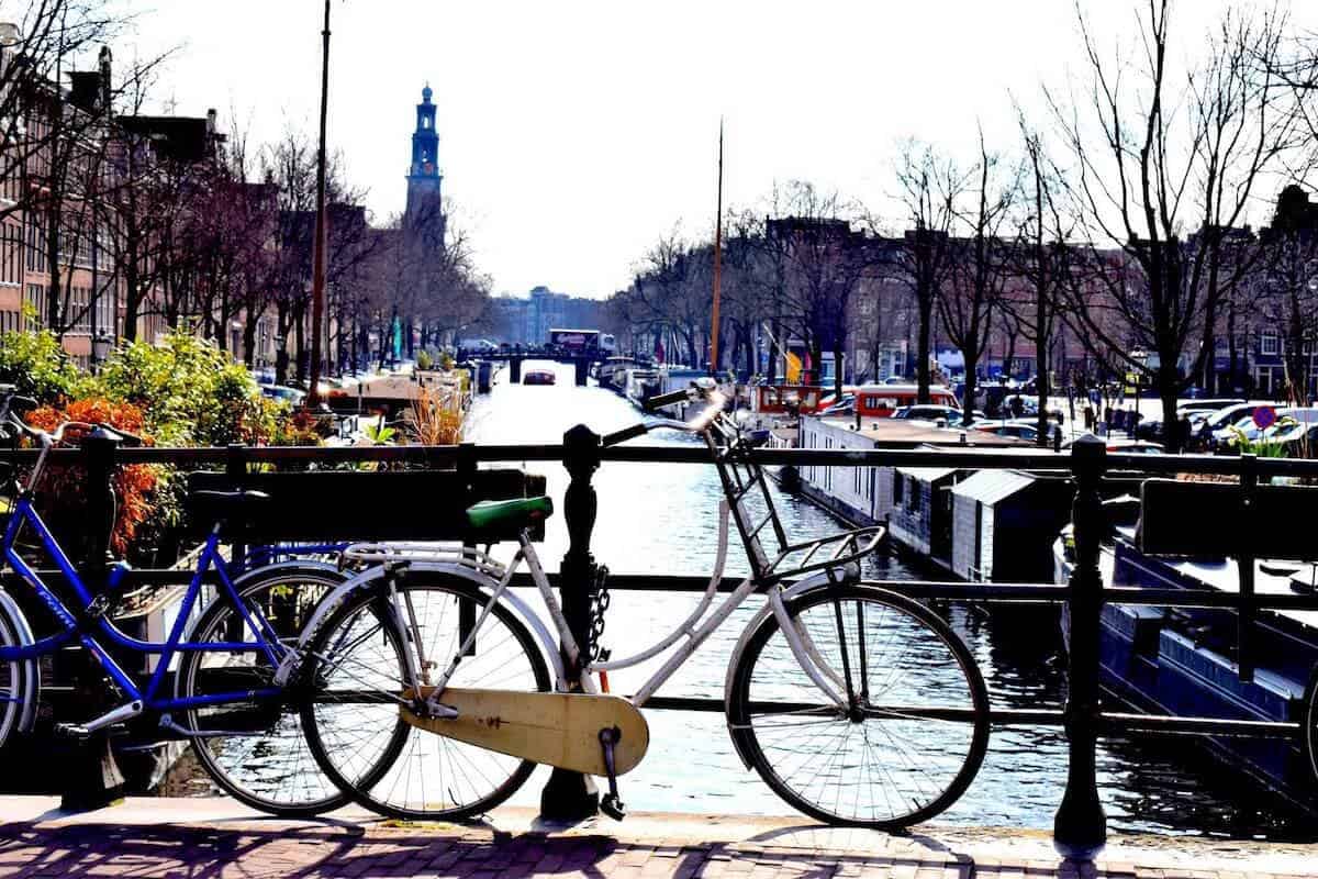 A white bicycle is locked to a black railing overlooking a canal in a city. The scene includes boats on the water, brick sidewalks, and a distant tower. The sky is clear, and the trees lining the canal are leafless.