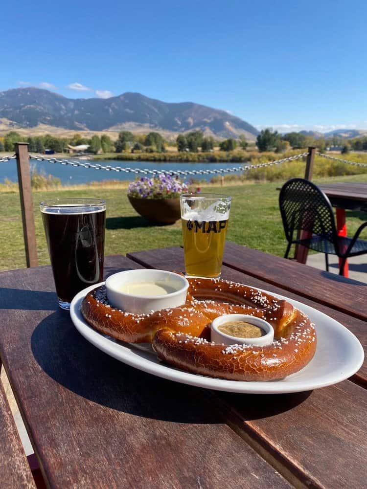 A large pretzel with dipping sauces on a white plate sits on a wooden table outdoors at MAP Brewing. Two glasses of beer are next to it. In the background, there's a view of a lake, mountains, and clear blue sky.