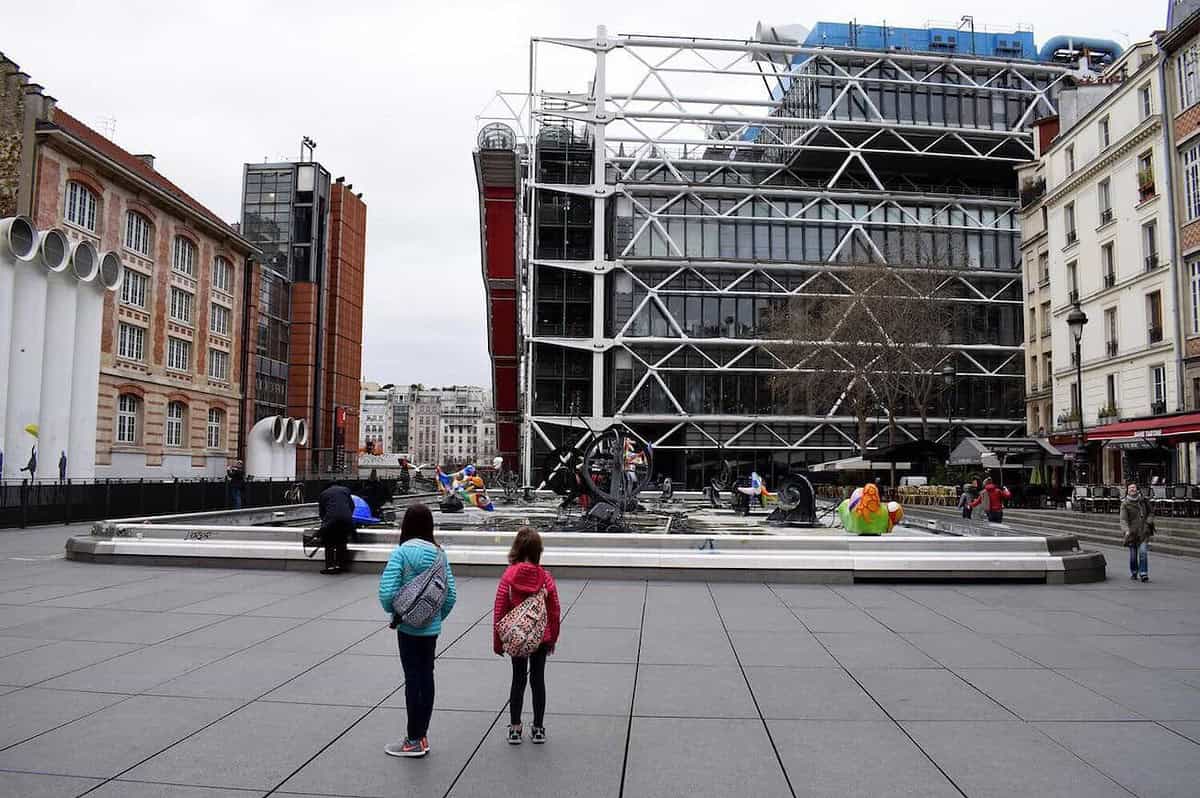 Two people stand in front of a modern building with an industrial design, featuring exposed pipes and metal framework. In the foreground, a fountain with colorful sculptures adds a playful touch, perfect for exploring Paris with kids. The surrounding area boasts a few classic-style buildings.