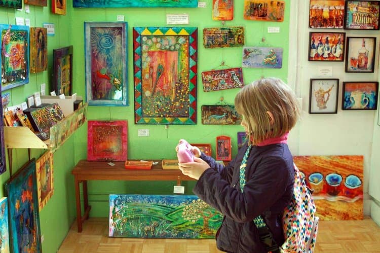 A child with a colorful backpack stands in an art gallery, observing a painting in a bright, eclectic room filled with vibrant artwork on green walls.