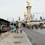 A family walks along a cobblestone street near the Seine River in Paris. The scene includes the ornate Pont Alexandre III bridge, with its golden sculptures and lampposts, and a riverside bistro in the background.