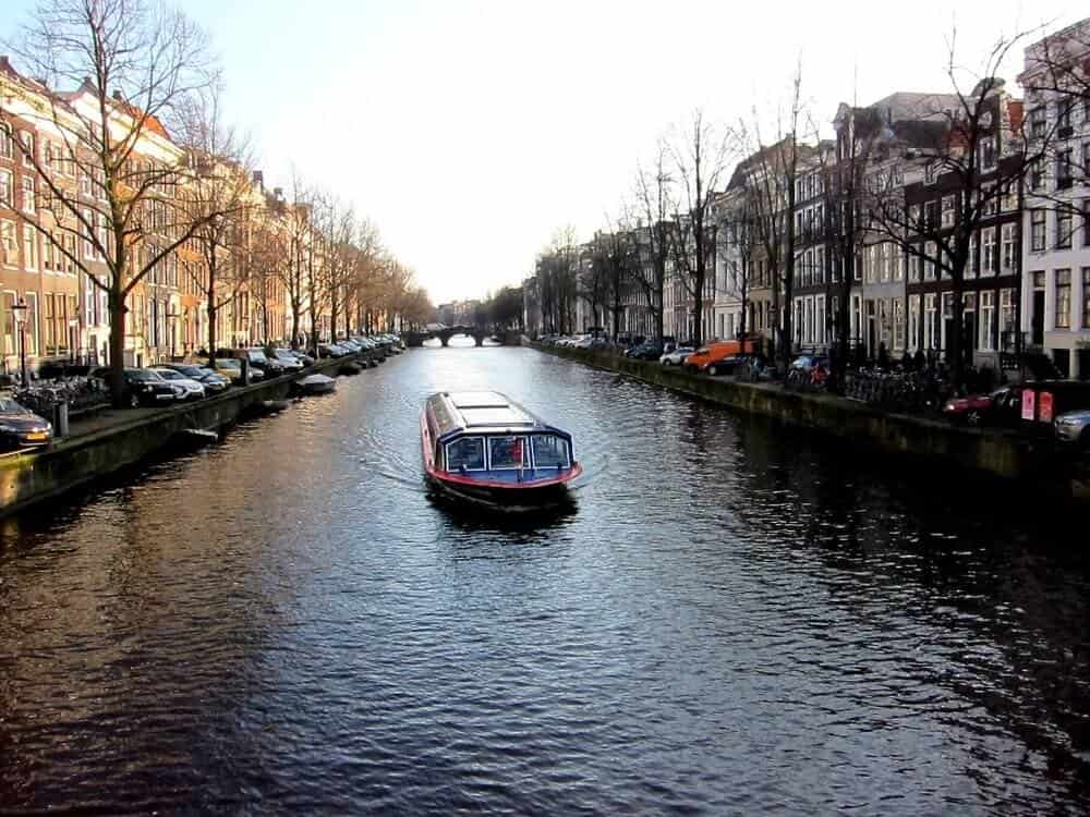 A canal boat cruises along a narrow waterway lined with parked cars and historic buildings, reflecting in the water under a clear sky. Leafless trees stand on both sides, suggesting a late autumn or early winter setting.