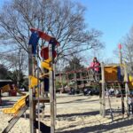 Children play on a colorful playground with climbing structures and slides in a park. The Eiffel Tower is visible in the background against a clear blue sky, and bare trees are scattered around the sandy area.