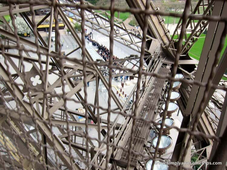 Explore Paris with kids as you view through the lattice framework of the Eiffel Tower, capturing geometric patterns and a glimpse of people waiting below. The lush greenery in the background contrasts with the iconic metal structure, making it a captivating sight for visitors of all ages.