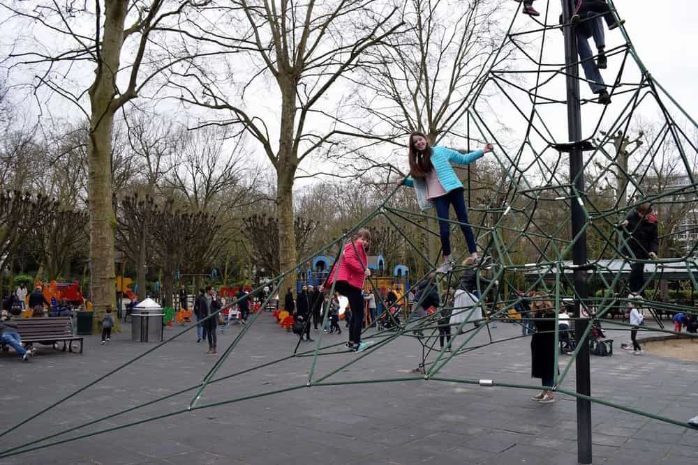 Children play on a geometric climbing structure in a park, capturing the charm of Paris with kids. One girl in a blue jacket climbs higher, while others are scattered below. Leafless trees surround the area as people stroll by on a cloudy day, creating an enchanting scene for families.