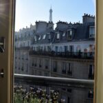 From an apartment window, the view stretches over Parisian buildings to the Eiffel Tower in the distance. Sunlight beams in, perfect for a family adventure exploring Paris with kids, highlighting a balcony flower box brimming with purple blooms.