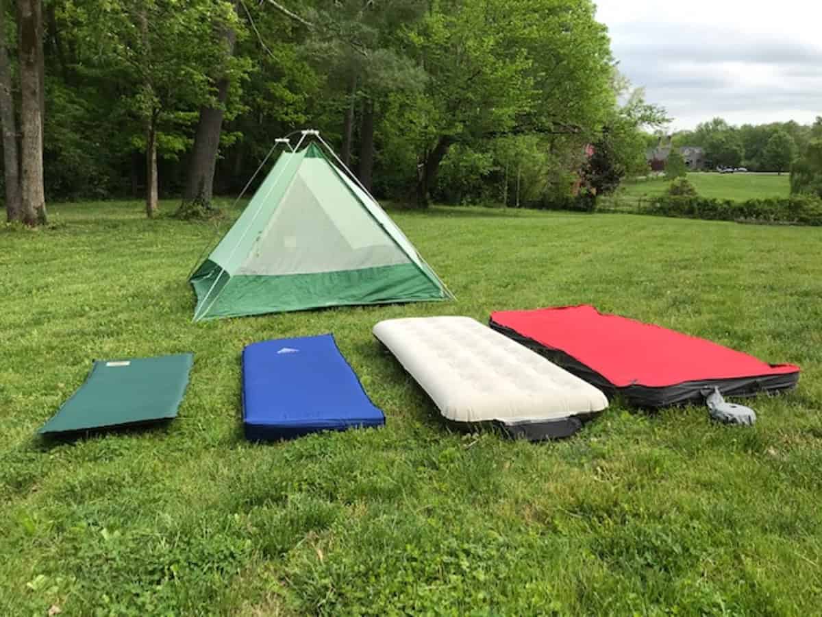 A small green tent is set up on a grassy field, surrounded by four different sleeping pads and mattresses, each laid out parallel to each other. Trees and a cloudy sky are visible in the background.