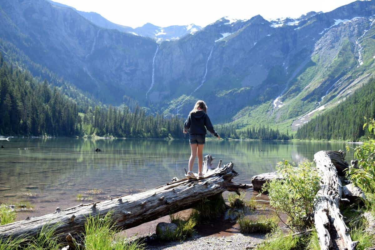 A person stands on a fallen log by a calm mountain lake, surrounded by evergreen trees and towering mountains. The sun illuminates patches of the landscape, highlighting distant waterfalls and rugged terrain.