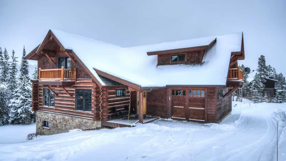 A snow-covered log cabin with a wooden balcony and stone foundation stands amid a snowy landscape. Surrounded by pine trees, the cabin has large windows and a sloped roof, creating a cozy winter scene.