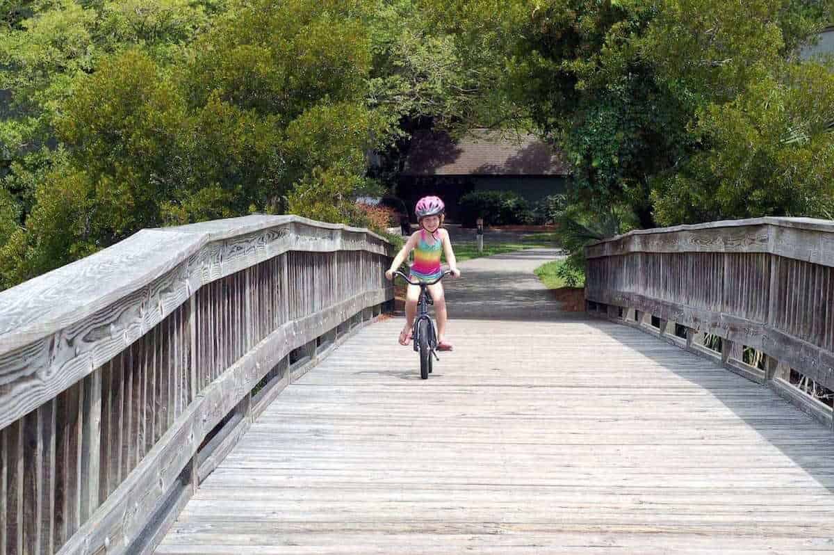A child wearing a colorful outfit and pink helmet rides a bicycle on a wooden bridge surrounded by lush green trees. The scene is bright and sunny.