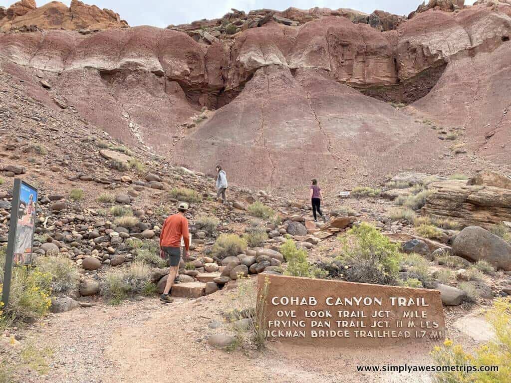 Three hikers walk on a rocky trail in front of a large red rock formation. A sign in the foreground reads, "Cohab Canyon Trail," with directions to other trails—a must-see among the things to do in Capitol Reef National Park. Sparse vegetation is visible in the rugged landscape.