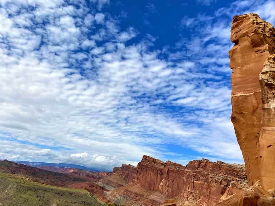 Rocky desert landscape with red sandstone cliffs under a bright blue sky dotted with white clouds—an iconic scene at Capitol Reef National Park. A large rock formation stands prominently on the right, while smaller cliffs stretch into the distance on the left. Discover endless things to do in this enchanting park.