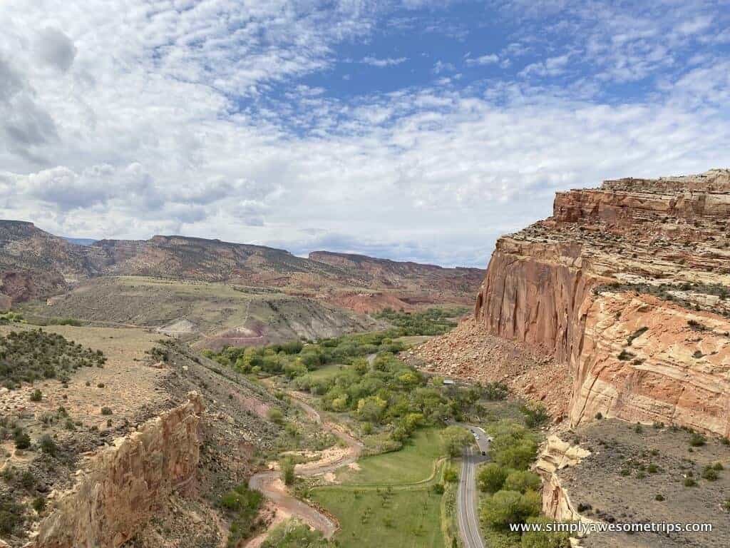 A scenic view of Capitol Reef National Park reveals a breathtaking canyon landscape with red rock cliffs, green trees, and a winding road below. The partly cloudy sky adds depth to this expansive setting, inviting exploration and adventure among the park’s many wonders.