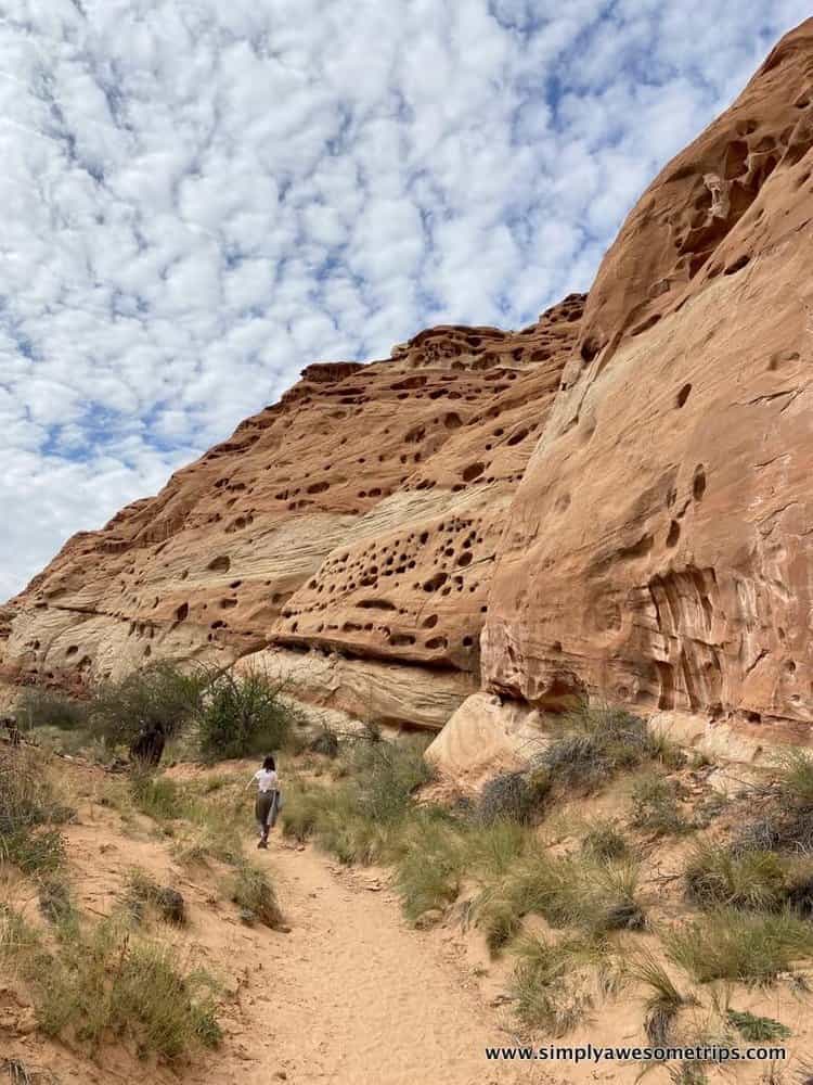 A person walks along a sandy trail beside a rugged, red rock formation with natural holes in Capitol Reef National Park. The sky is partly cloudy, and there is sparse vegetation along the path. Exploring this unique landscape is one of the best things to do in the park.