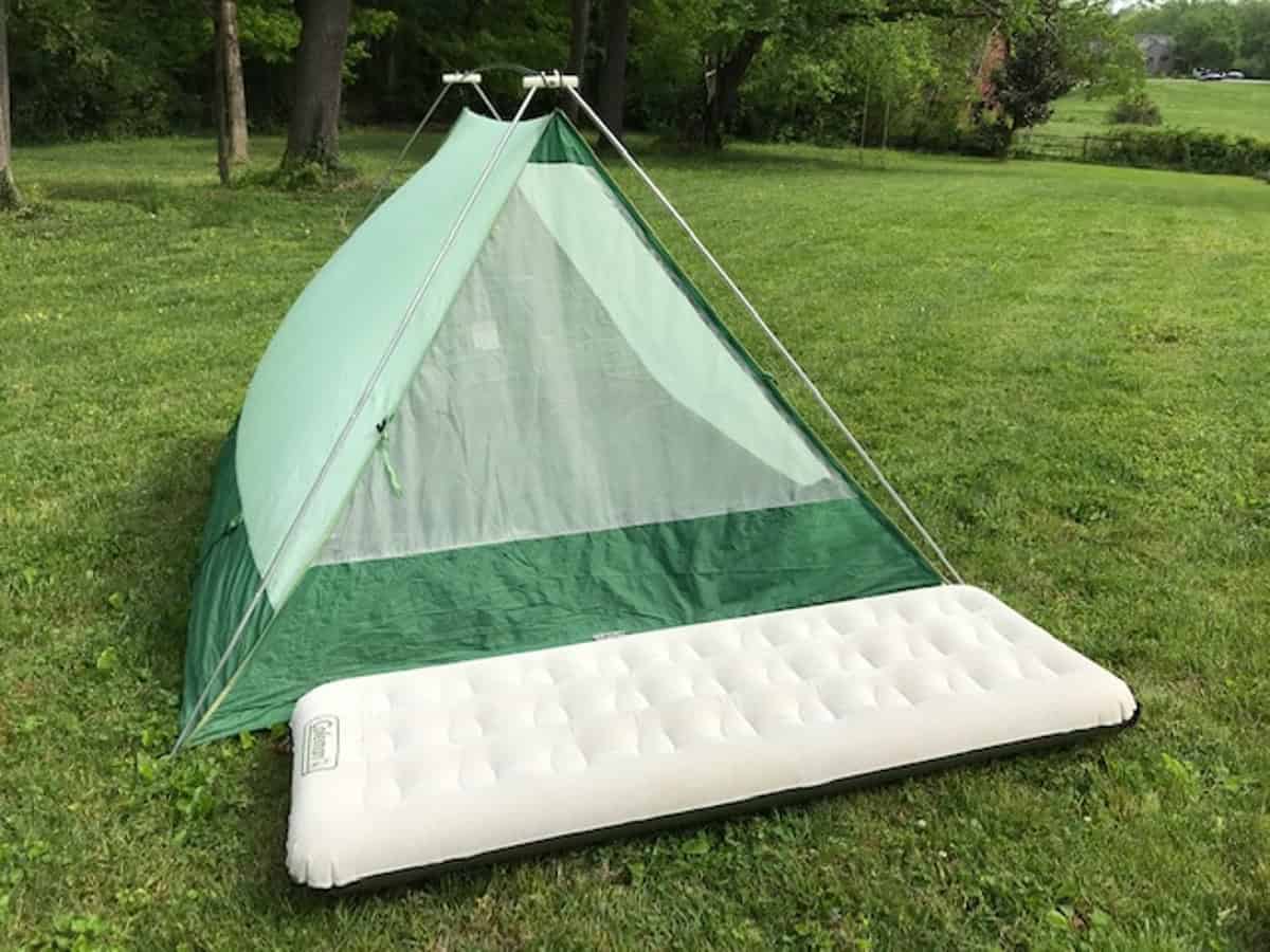A green tent is set up on a grassy field with a white air mattress placed in front. Trees and a distant landscape are visible in the background under a clear sky.