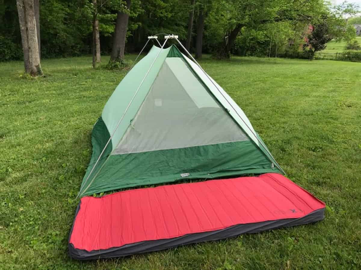 A green and gray camping tent set up on a grassy lawn with a red sleeping pad laid out in front. Tall trees and lush greenery surround the area, creating a serene outdoor setting.