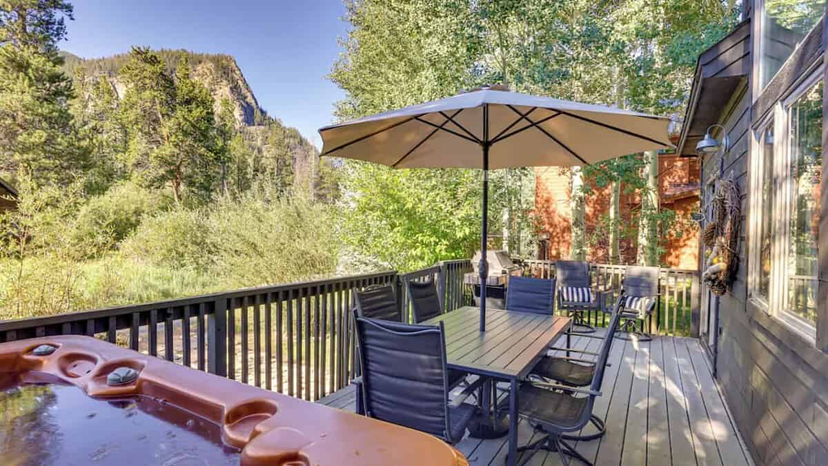 Outdoor deck with table, chairs, and an umbrella. Surrounded by trees and mountains in the background. A hot tub is in the foreground, and the scene is bathed in sunlight from a clear blue sky.