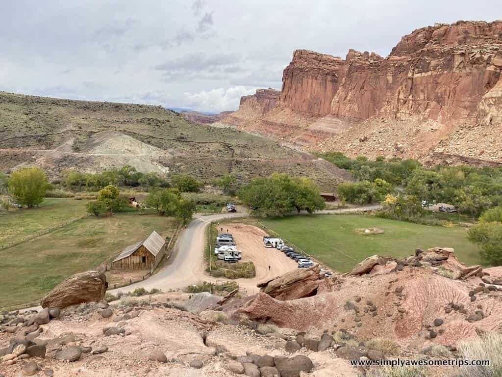 A scenic view of Capitol Reef National Park showcases red rock cliffs, a grassy area with trees, and a rustic wooden cabin. Parked cars line the dirt road as visitors explore things to do in Capitol Reef National Park under a cloudy sky, capturing an expansive and serene landscape.