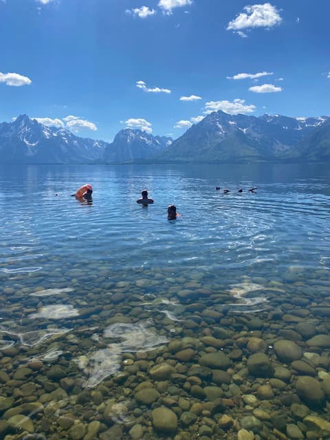 People swimming in clear, shallow lake water against a backdrop of snow-capped mountains and a blue, cloud-dotted sky. The rocky lakebed is visible under the water.