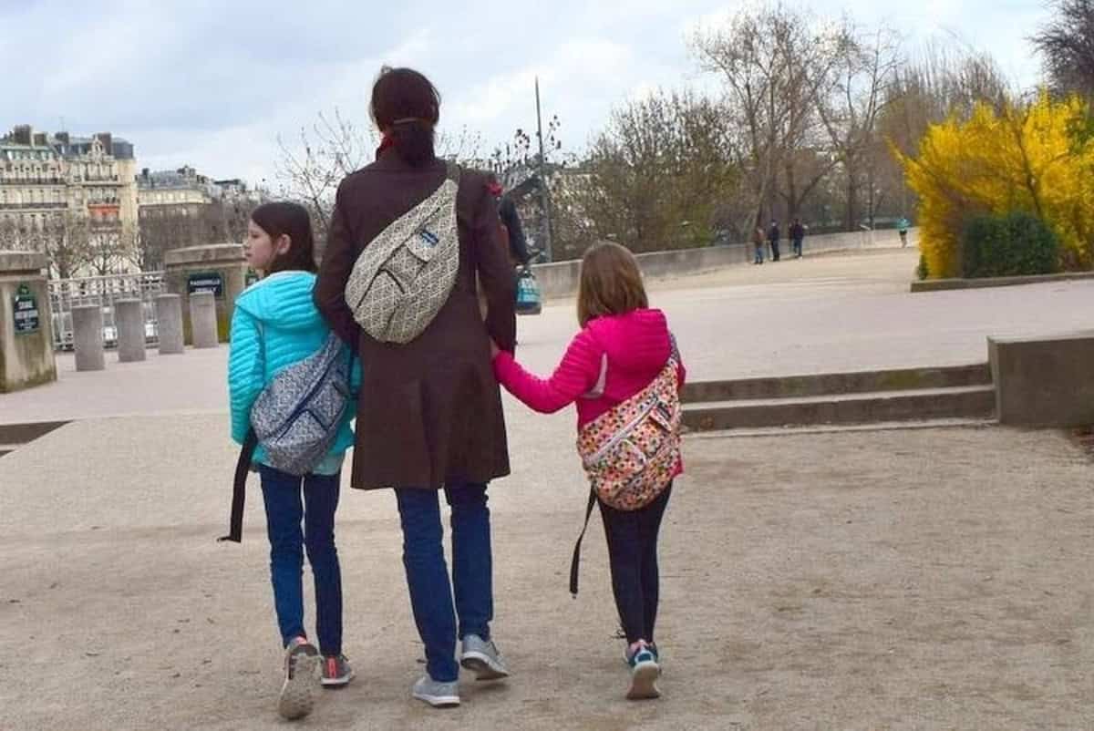 A woman walking with two children through a park. The woman is in the center, wearing a coat and carrying a sling bag. The children, each wearing bright jackets, walk on either side, holding hands. They all have patterned backpacks. Trees and buildings are visible in the background.