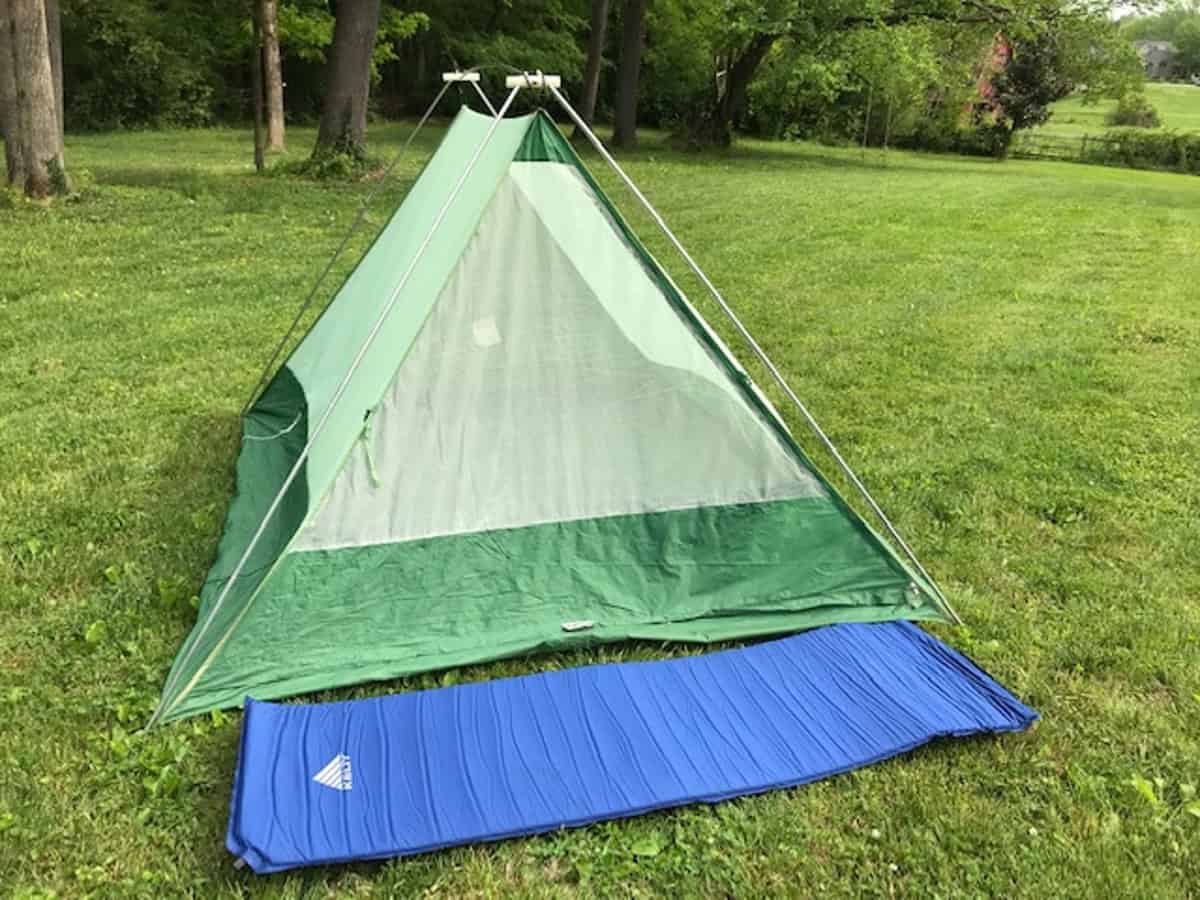 A green triangular tent set up on a grassy area, with forest trees in the background. A blue sleeping mat is laid out in front of the tent.