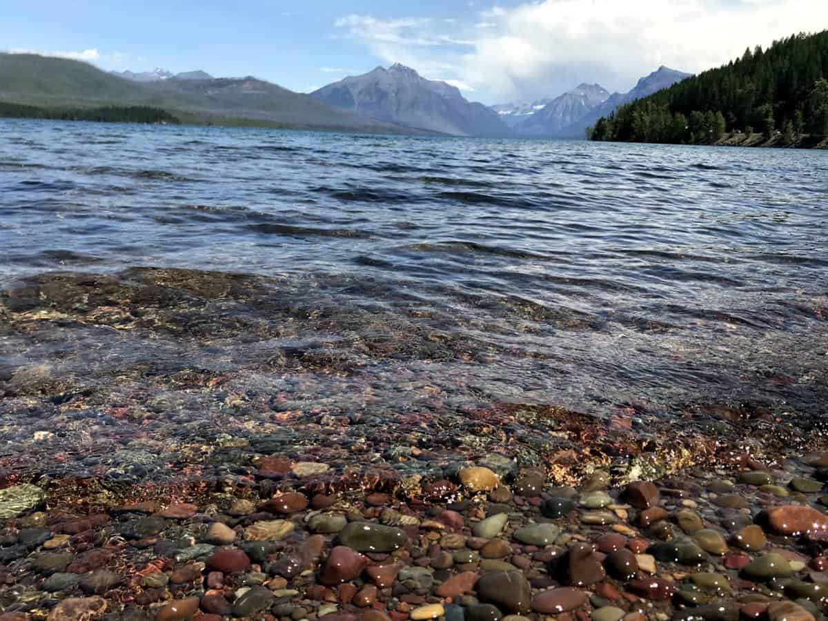 A scenic view of a lake with clear, shallow water revealing colorful pebbles. In the background, rugged mountains rise under a partly cloudy sky. Dense evergreen forests line the distant shoreline.