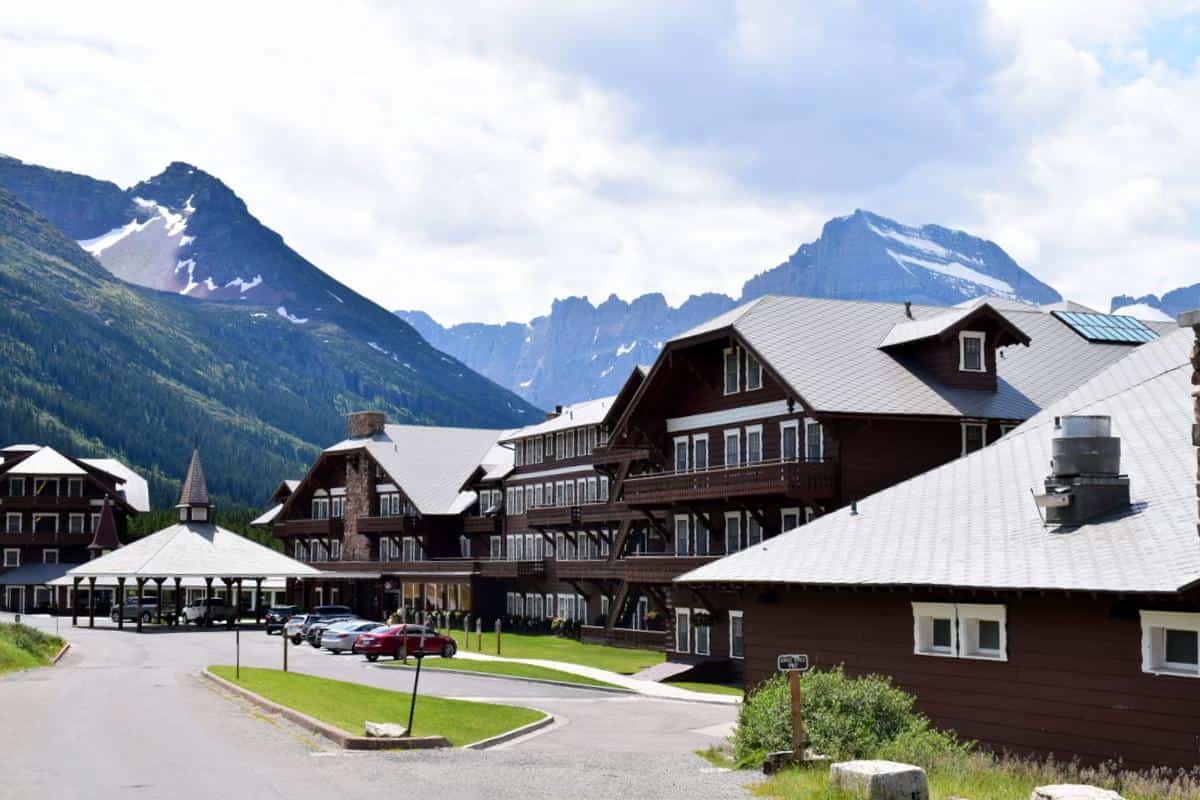 A rustic lodge with dark wood exterior nestled in a picturesque mountain setting. Snow-capped peaks and lush greenery surround the buildings. Cars are parked on a paved path, and the sky is partly cloudy.