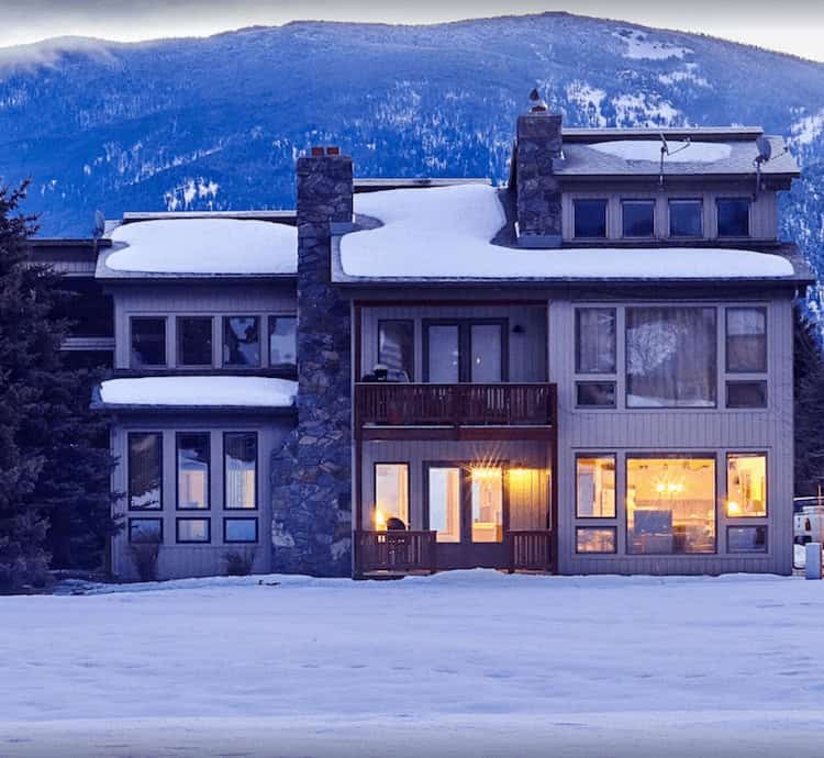 A large house with a stone chimney and large windows, warmly lit from inside, sits amidst a snowy landscape. In the background, snow-covered mountains rise against a dusky blue sky.