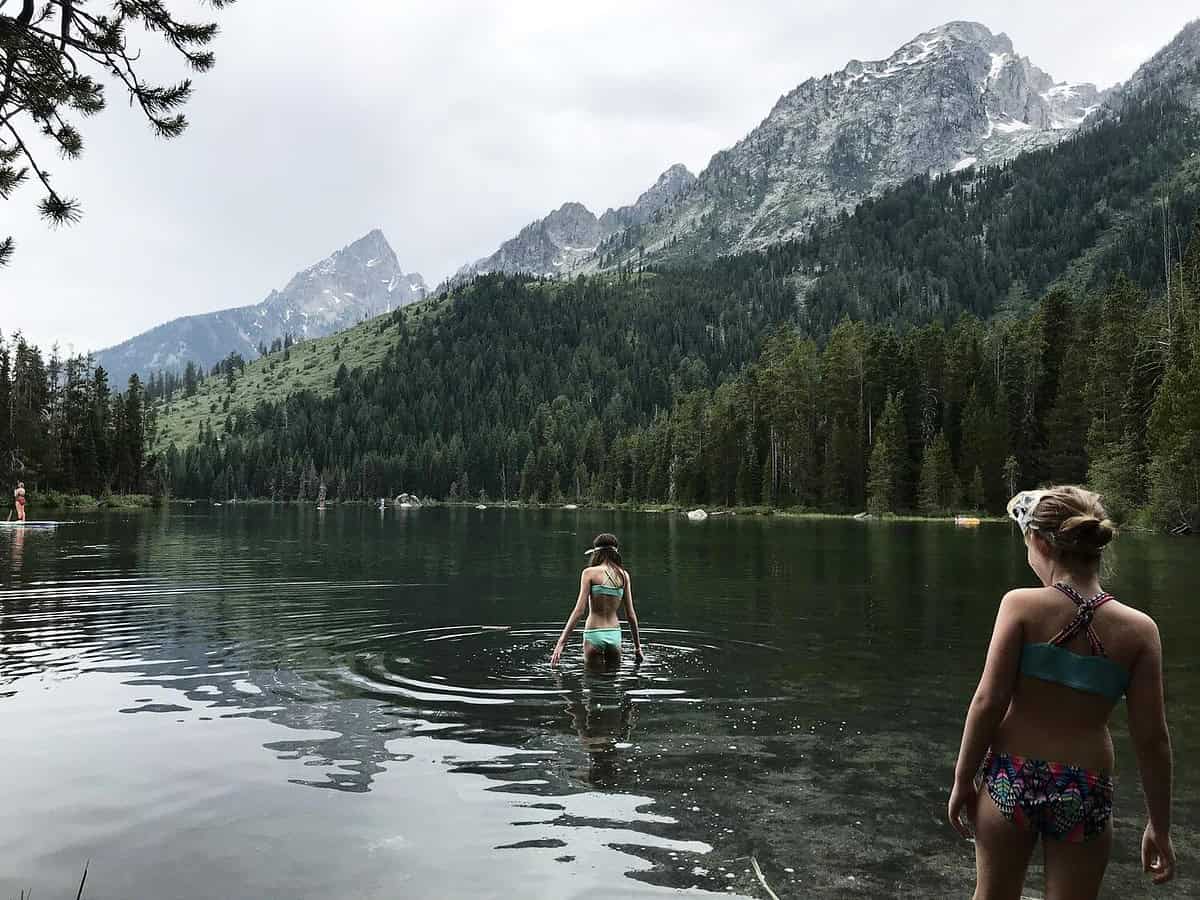 Two people in swimwear wade into a calm lake surrounded by dense pine forests and towering mountains. The scene is tranquil, with reflections on the water and overcast skies adding to the serene atmosphere.