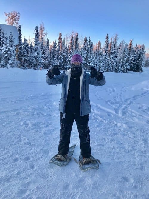 A person stands on a snowy landscape wearing snowshoes, a blue jacket, black pants, and a purple hat. Snow-covered trees are visible in the background under a clear sky. The person is pointing both thumbs up.