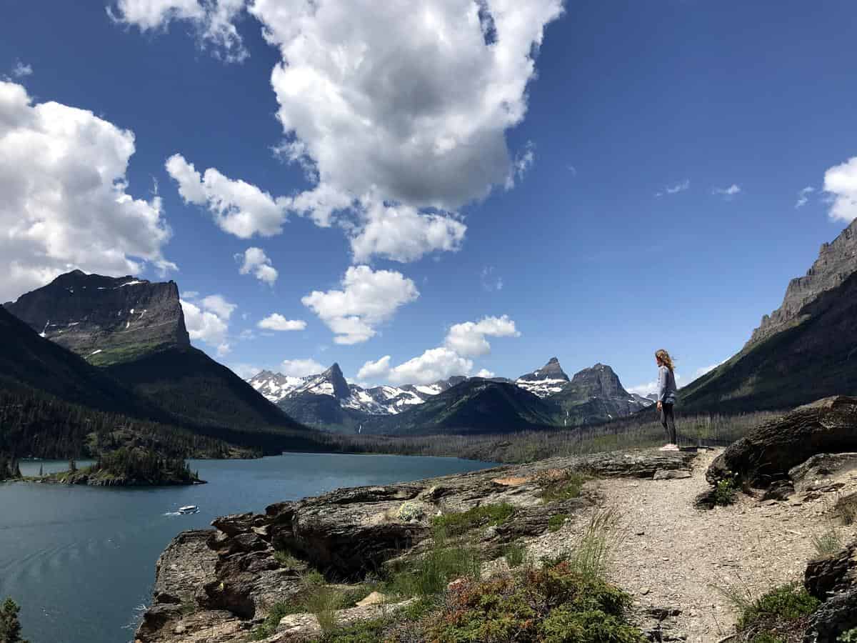 A person stands on a rocky cliff overlooking a scenic landscape with a large lake, surrounded by mountains under a partly cloudy sky. Snow-capped peaks are visible in the distance, and lush greenery covers the area.