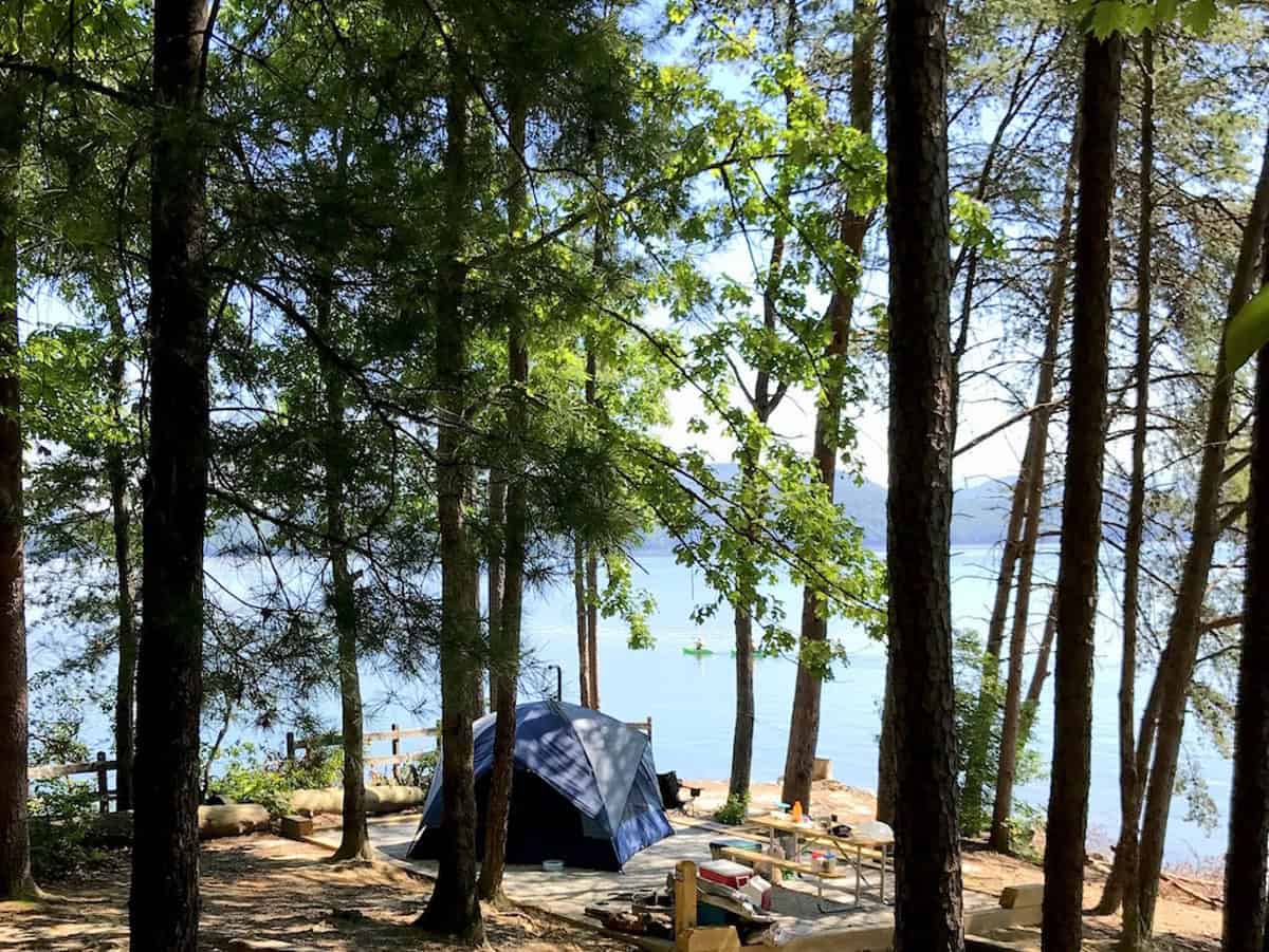 A blue tent is set up in a forested area overlooking a tranquil lake. Sunlight filters through the tall trees, illuminating a picnic table and scattered camping gear nearby. The scene captures the serenity of nature and outdoor camping.