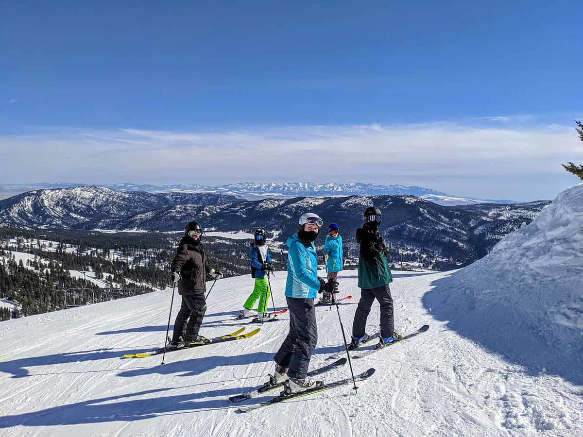 Five skiers stand at the top of a snowy mountain slope, wearing colorful ski gear. The expansive view shows snow-covered mountains under a clear blue sky in the background.