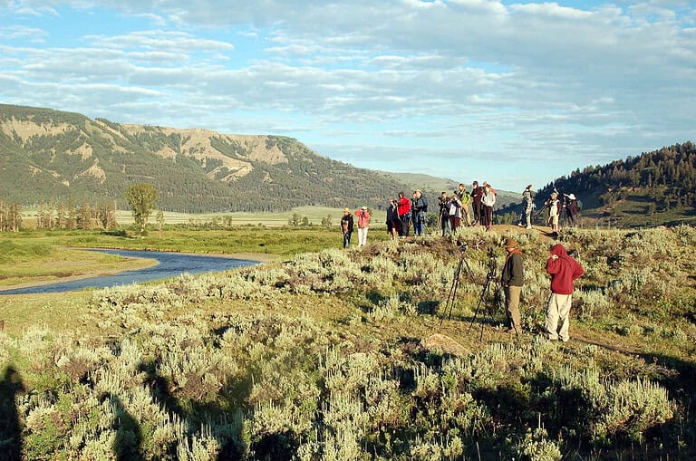A group of people stand on a grassy hill observing through binoculars and cameras. A winding river flows nearby, with mountains and trees in the background under a partly cloudy sky.