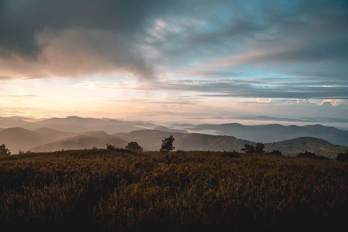 A scenic view of rolling hills and mountains covered in green vegetation under a cloudy sky, perfect for biking the Swamp Rabbit Trail with kids. The sun casts a warm, golden light over the landscape, creating a tranquil and serene atmosphere.