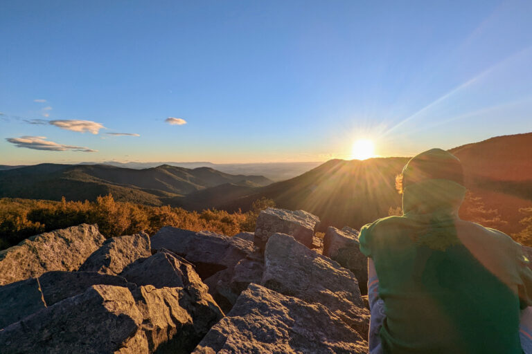 A person in a hoodie sits on rocky terrain, facing a sunset over a range of forested mountains. The sky is clear blue with a few clouds, creating a peaceful and scenic vista over Shenandoah National Park.