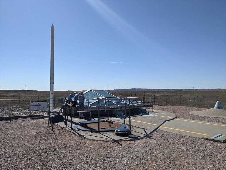 A group of people stand around a glass enclosure at a missile launch site on a clear day. The enclosure is surrounded by a fence, and a tall, thin structure is nearby. The landscape is flat with sparse vegetation.