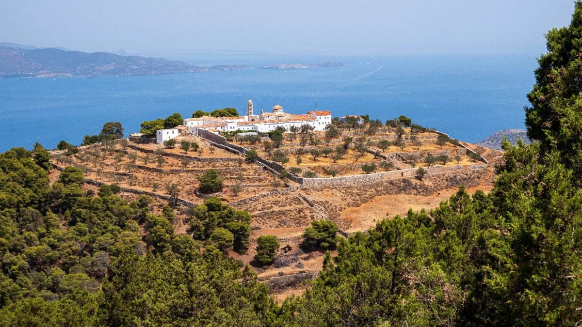 A hilltop monastery surrounded by dry, terraced land and trees overlooks a blue sea. The buildings have white walls and red roofs. The coastline and distant hills are visible in the background under a clear sky.