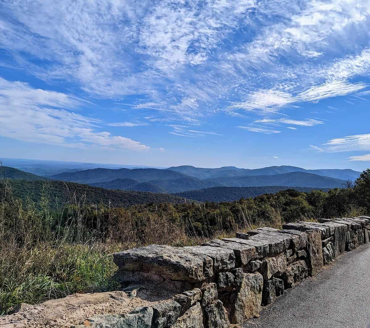 A scenic view from a stone wall-lined overlook, showcasing rolling mountains under a clear blue sky with scattered clouds. Grass and shrubs are visible in the foreground, enhancing the natural beauty of the landscape—perfect for daydreaming about your next adventure with airport lounge access for the family.