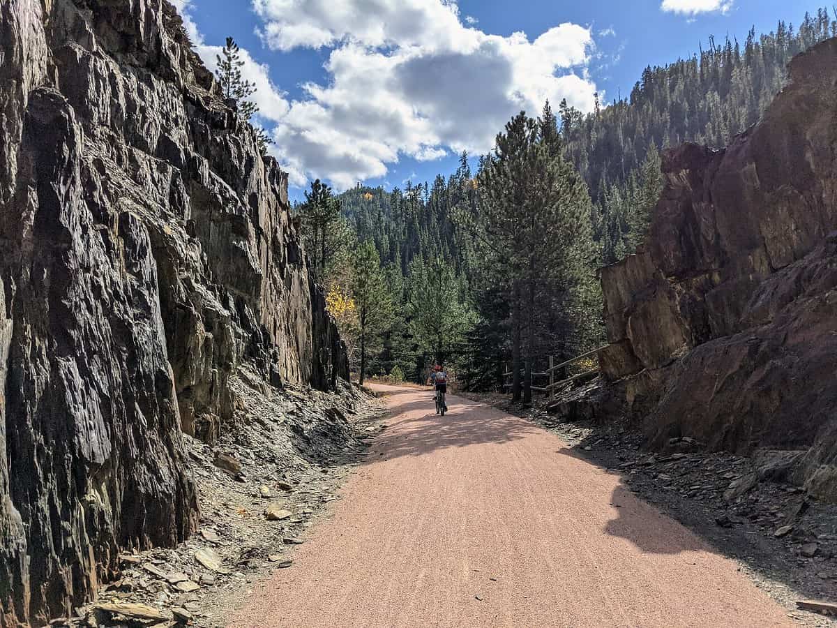 A person walks a bicycle down a dirt path through a forested mountain area. Cliffs rise on both sides of the path, with pine trees and a blue sky with fluffy clouds above.