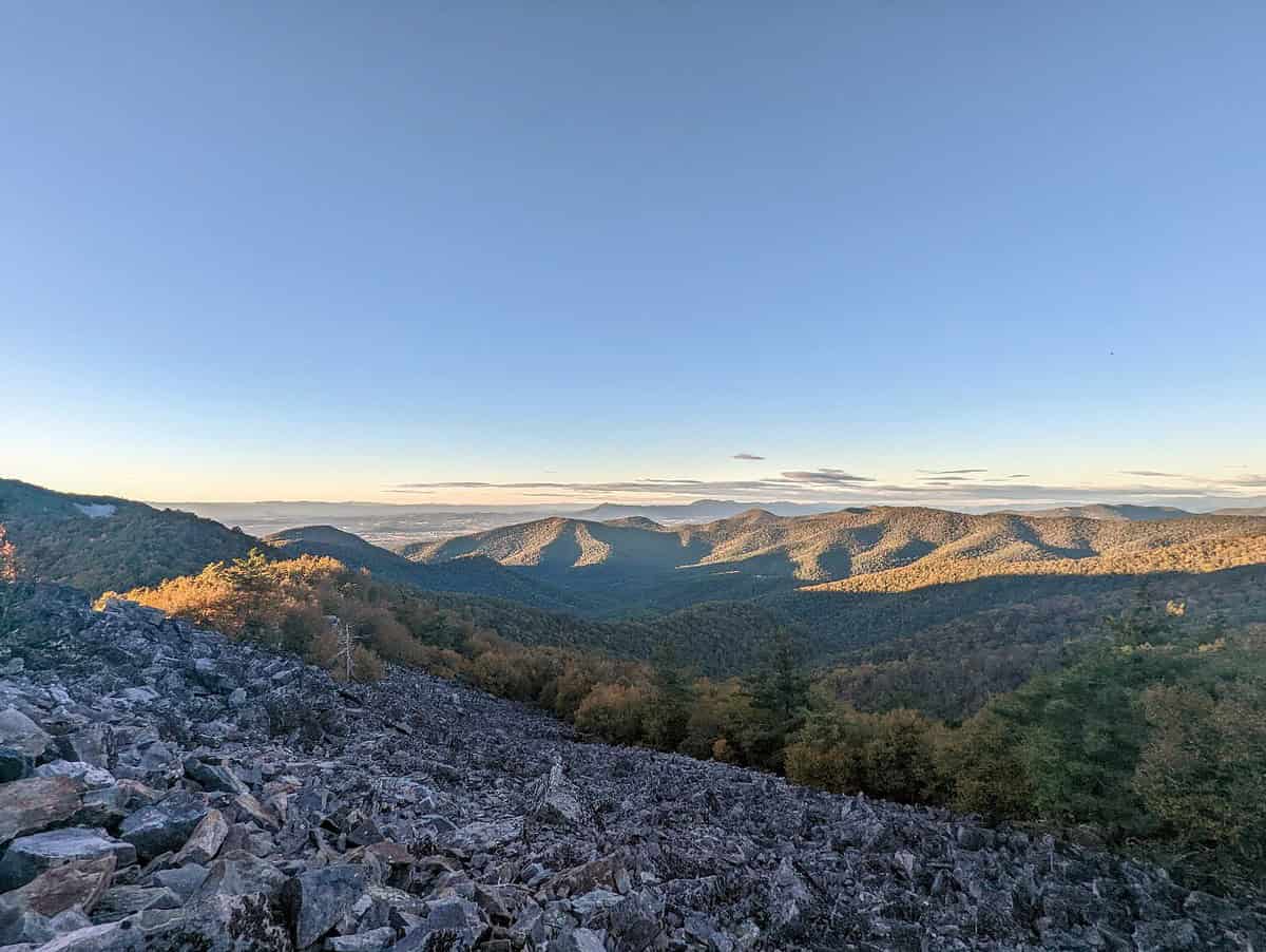 A vast landscape view of rolling green mountains under a clear blue sky during sunset. The foreground features rocky terrain, with sunlight casting a warm glow on the hills and trees, creating a serene and picturesque scene.