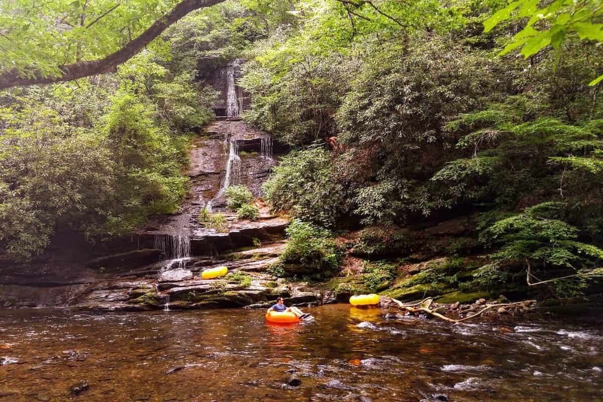 People are tubing in a serene forest setting, floating on inflatable tubes in a clear river with lush green foliage surrounding them. Nearby, families are biking the Swamp Rabbit Trail with kids. In the background, a gentle waterfall cascades down a rock face.