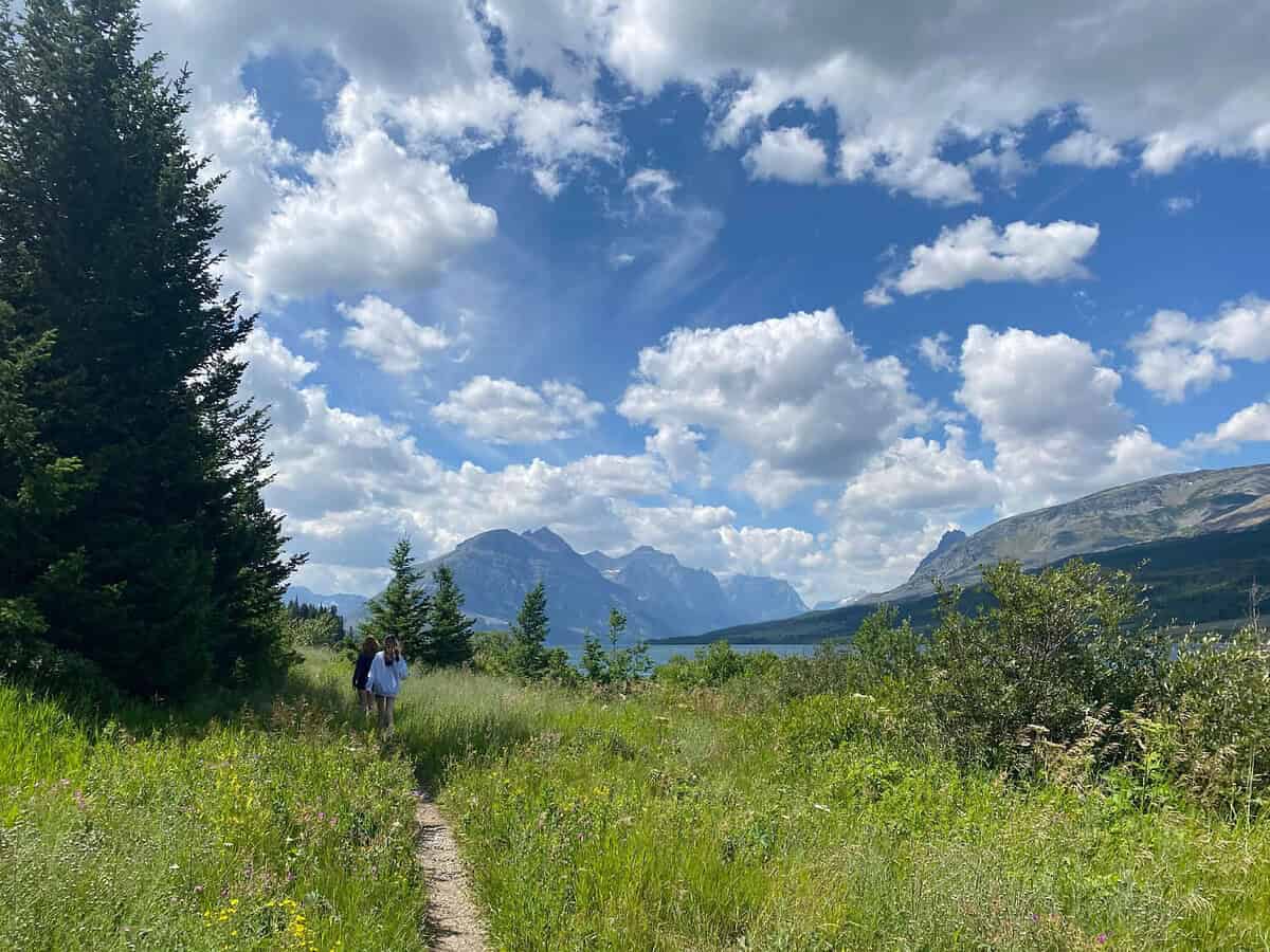 Two people walk on a narrow dirt trail through tall grass and wildflowers, surrounded by trees and mountains under a partly cloudy sky.