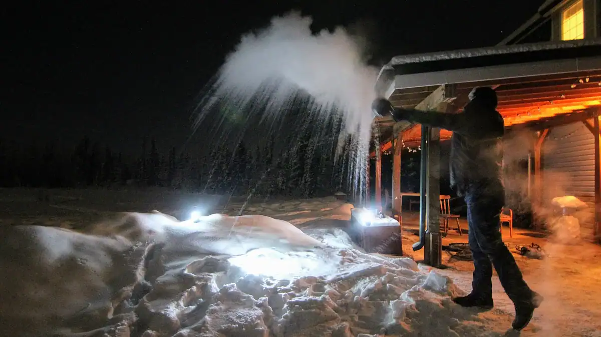 A person outside a house on a snowy night throws water into the air, creating a cloud of steam as it freezes in the cold. Bright lights illuminate the snow and wooden porch.