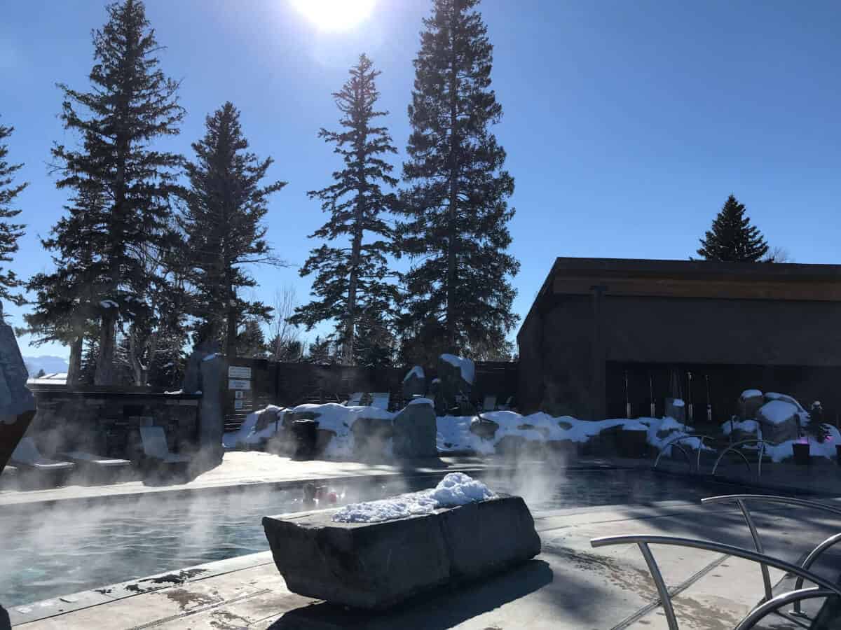 A steaming outdoor pool in a snowy mountain setting, surrounded by pine trees and a wooden building.